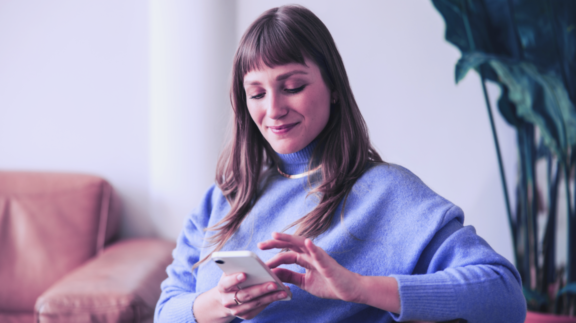 A woman wearing a blue sweater sits indoors and smiles while using her smartphone, with a couch and large plant visible in the background.