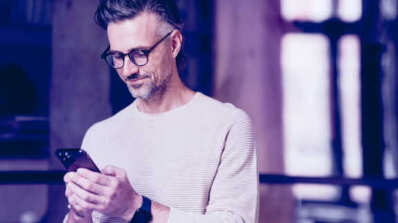 A man with glasses focuses on his phone, appearing engaged with the content displayed on the screen.