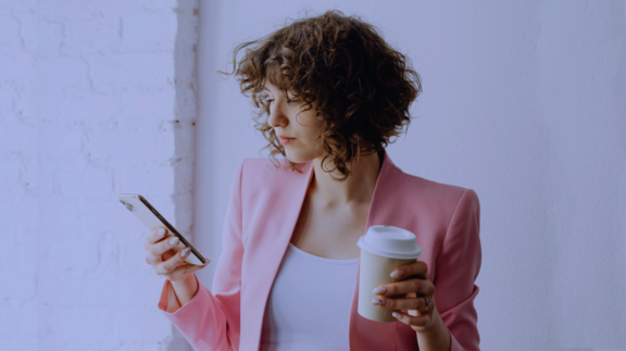 A person with short, curly hair wearing a pink blazer and holding a smartphone in one hand and a disposable coffee cup in the other. They appear to be focused on the phone. The background is minimalistic, featuring a white brick wall and natural lighting.